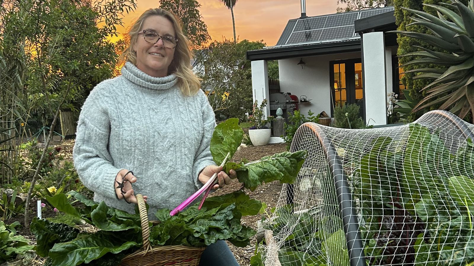 A smiling woman in a grey knit sweater holds a wicker basket of chard and a few pink-stemmed stalks in a garden at sunset, with a white house and a palm behind her