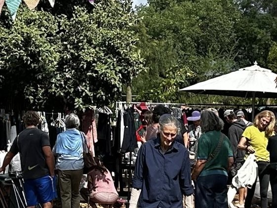 Shoppers browsing stalls under trees and bunting at a seasonal market in the garden, with a woman carrying a woven basket in the foreground
