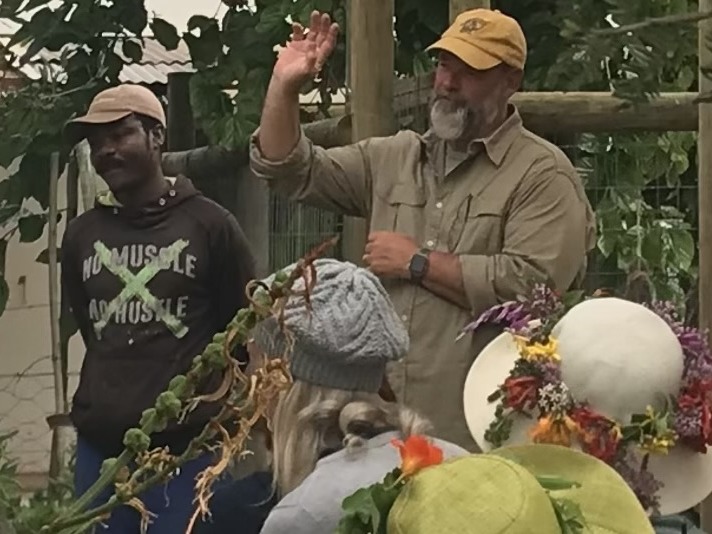 Two presenters talking to a workshop group in the garden, with a flower-crowned sun hat in the audience